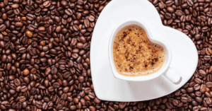With a background of roasted coffee beans sits a white heart shaped cup and saucer full of hot coffee, representing the thoughtful kindness of a gift from Innovation Coffee.