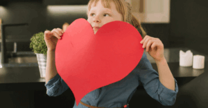 A child holds a large red paper heart, peaking over the top, representing acts of kindness