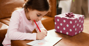 A girl used a red pencil crayon to write birthday messages inside a card. She is using a coffee table to write at, while sitting on the floor. There is a wrapped gift on the table beside the card.