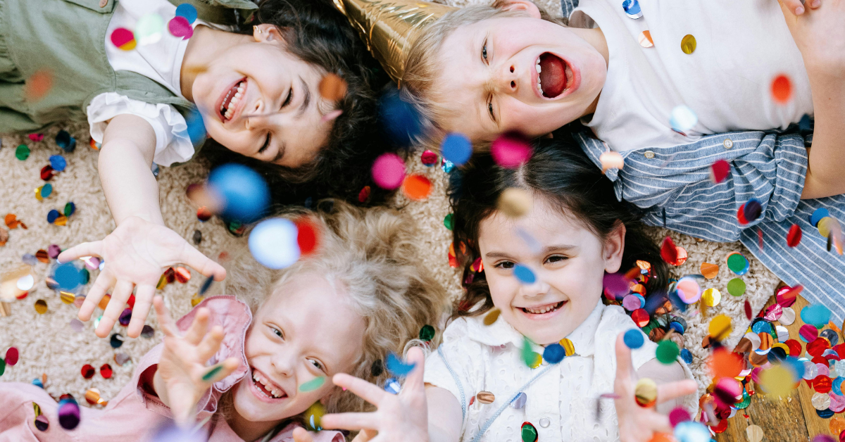 4 happy children, laying on the floor looking up towards the overhead camera, huge smiles of delight, colourful confetti showering down on them, clearly a magical birthday moment.