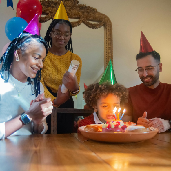 A typical scene in Canadian families celebrating birthdays, a child blows out candles, which are on a birthday donut at a table with their mother and father and big sister, who all wear shiny party hats. Everyone is smiling and there are balloons in the background.