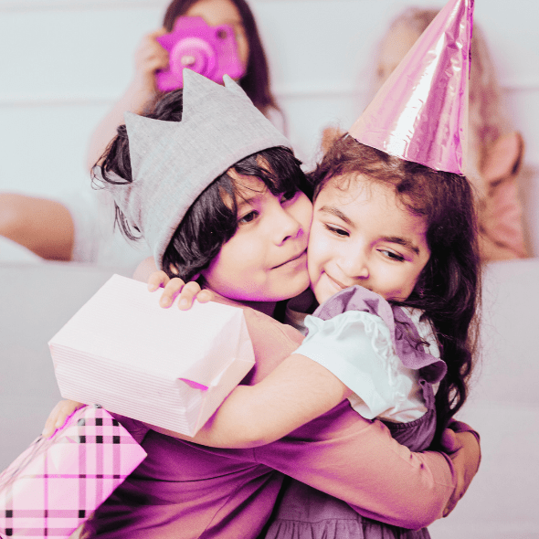 A child wearing a grey felt crown and a child wearing a shiny birthday hat and lavender dress share a hug, while holding wrapped gift boxes. Someone in the background is holding a camera to take a picture.