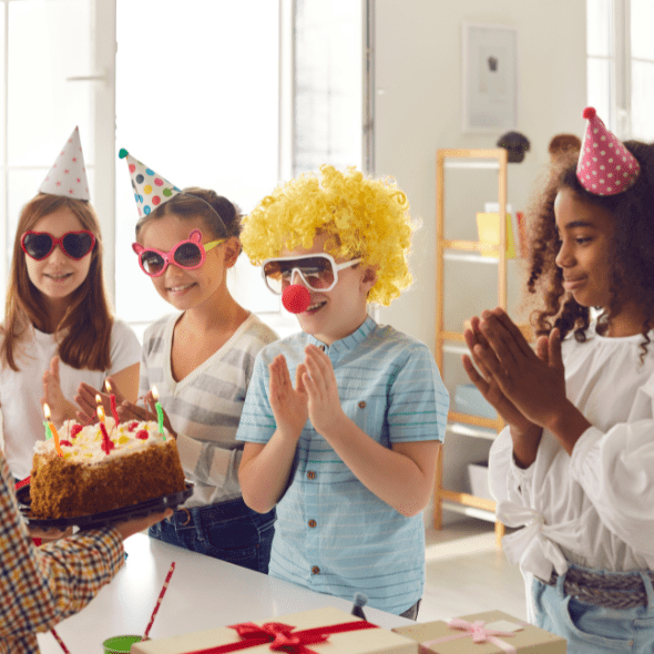 Children celebrating a birthday part at home, wearing sunglasses, wigs, and party hats. The birthday child is being handed a slice of chocolate cake.
