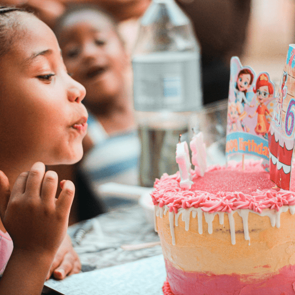 A girl blows out her birthday candles as her brother looks on, smiling in the background. The background is blurred, to focus on the birthday girl and her pink cake decorated with princesses and candles.