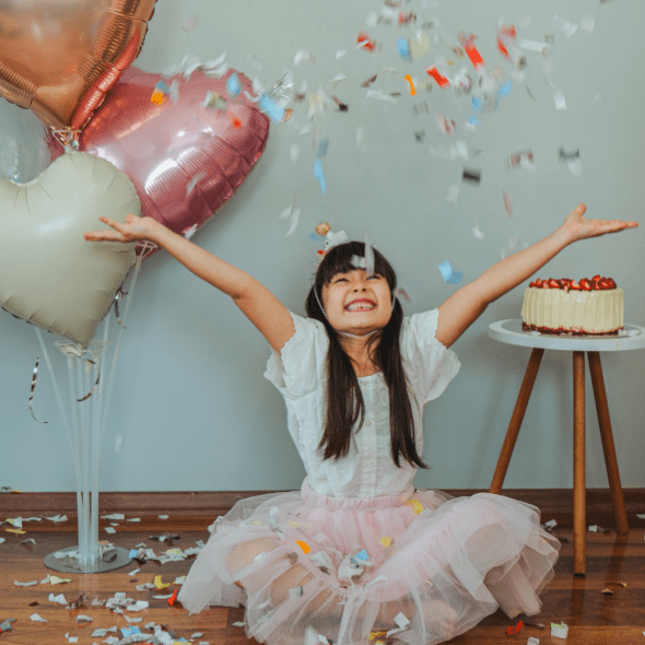 A child wearing a pink frilly dress and white shirt with puffy sleeves sits on the floor, happily tossing confetti into the air, balloons and birthday cake behind her off to the sides.