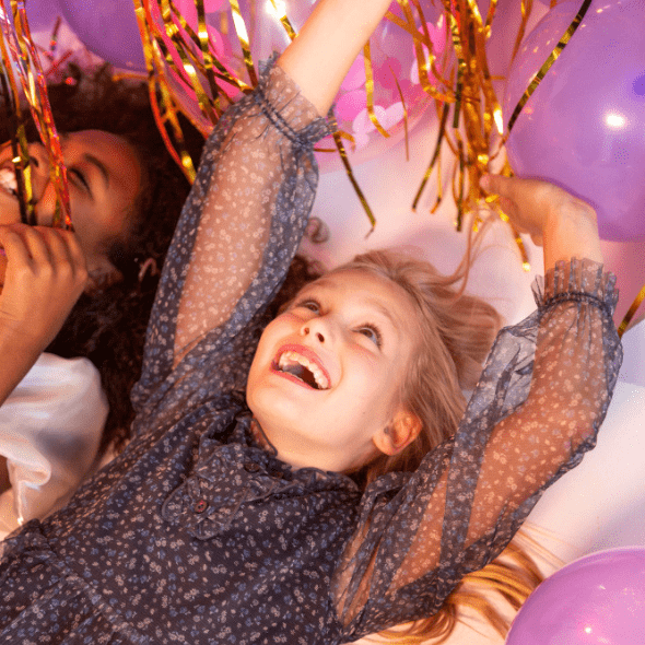 Two children in a moment of joy, lay on their backs next to each other, delighting in the purple birthday balloons and gold streamers.