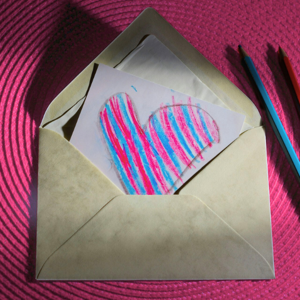 An open card envelope sits open on a pink circular placemat, next to blue and pink pencil crayons. Inside the envelope is a hand drawn heart that is striped pink and blue, that extend beyond the uneven outline.
