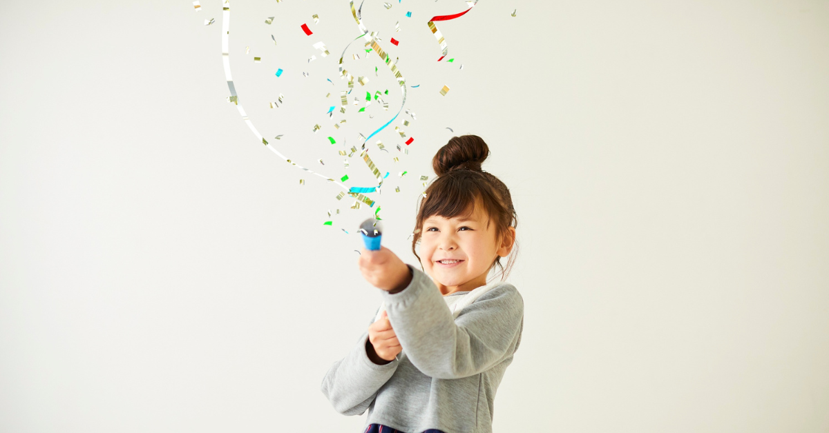 An Asian girl shoots a confetti blaster into the air, grinning as the colourful pieces fly upward. She is positioned on a simple white background and wears a plain grey sweater with her hair in a messy bun lopsided on the top of her head.
