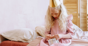 A girl with long blond hair wearing a pink dress and gold party had sits on her bed opening a small gift. Why birthdays matter for kids is about belonging and feeling valued.